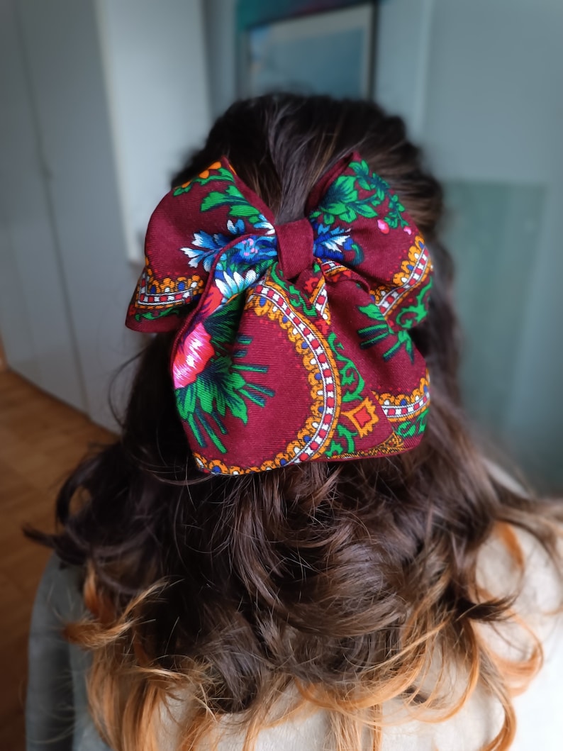 May include: A large burgundy bow with a floral pattern in shades of green, blue, red, and yellow. The bow is tied in a large, full knot and is worn in the hair.
