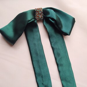 May include: A dark green satin bow with a beaded embellishment in the center. The bow is tied with two long tails.