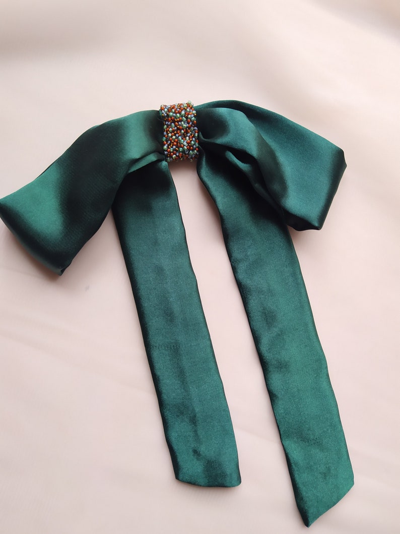 May include: A dark green satin bow with a beaded embellishment in the center. The bow is tied with two long tails.