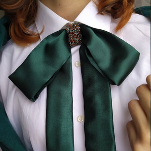 May include: A dark green satin bow tie with a small, beaded embellishment in the center. The bow tie is tied around a white collared shirt.