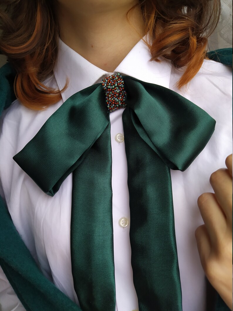 May include: A dark green satin bow tie with a small, beaded embellishment in the center. The bow tie is tied around a white collared shirt.