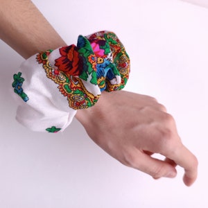 May include: White fabric scrunchies with colorful floral patterns. The scrunchies feature red, blue, green, and yellow flowers. The scrunchies are a hair accessory.