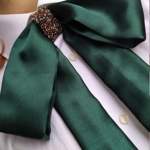 May include: A dark green satin bow tie with a beaded embellishment. The bow tie is tied around a white collared shirt.