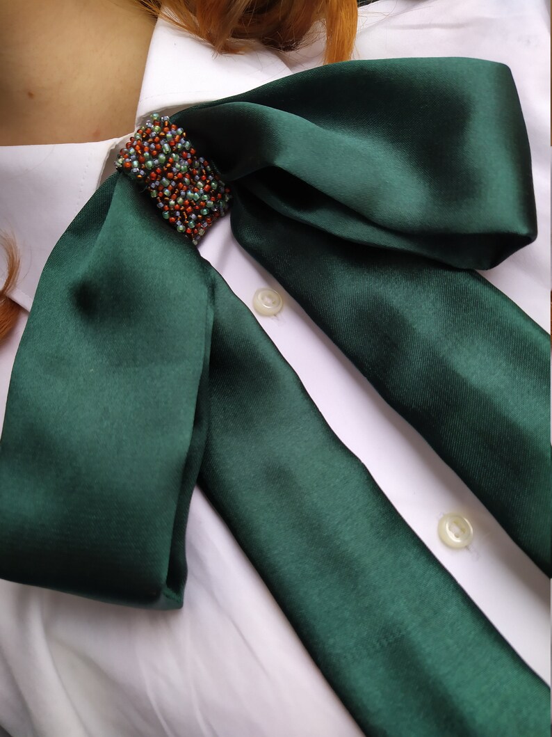 May include: A dark green satin bow tie with a beaded embellishment. The bow tie is tied around a white collared shirt.