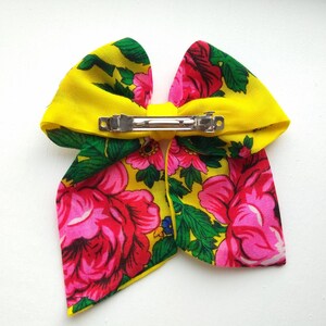 May include: A yellow fabric bow with a floral pattern in pink, green and blue. The bow has a silver metal clip on the back.