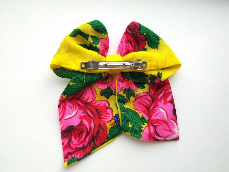 May include: A yellow fabric bow with a floral pattern in pink, green and blue. The bow has a silver metal clip on the back.