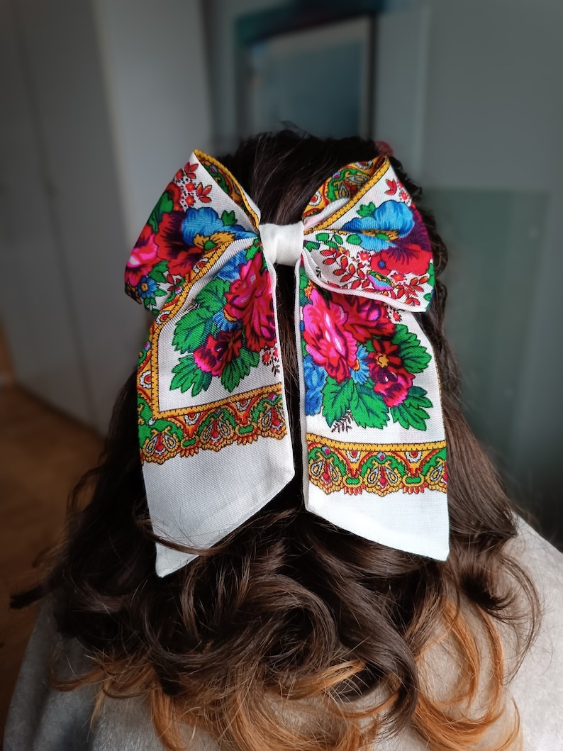May include: A white bow with a floral pattern in shades of red, pink, blue, green, and yellow. The bow is tied in a large knot and is made of a soft, lightweight fabric.