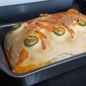 May include: A loaf of bread topped with melted cheese and green jalapeno peppers in a gray baking dish.
