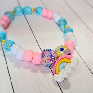 May include: A colorful beaded bracelet with a rainbow and cloud charm. The bracelet is made with pink, white, and blue beads, and the charm is made with a yellow, orange, red, blue, and green rainbow and a white cloud.