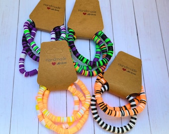 May include: Several colorful bracelets with a flat, disc-like design are displayed. The bracelets feature various color combinations, including purple, green, black, orange, and white. Each bracelet is attached to a brown tag with the text "Handmade with love."