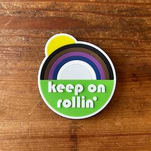 May include: A green, brown, purple, blue, and white rainbow with a yellow sun on top. The text "keep on rollin'" is written in white on the green background.