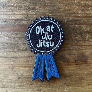 May include: A blue and white embroidered patch with a black background and the text "Ok at Jiu Jitsu". The patch has a blue ribbon attached to the bottom.