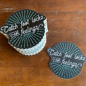May include: A stack of black and white stickers with a blue design. The stickers say "Catch foot locks, not feelings."