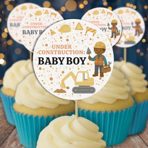 May include: Cupcakes with light yellow frosting in blue liners, topped with circular toppers. The toppers feature a construction theme with the text "UNDER CONSTRUCTION: BABY BOY" and illustrations of a construction worker, tools, and a yellow excavator.