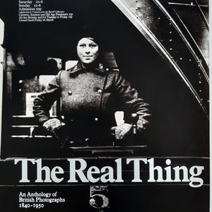 May include: Monochrome poster advertising "The Real Thing," an anthology of British photographs from 1840-1950. The image shows a person in a coat. Text includes "Hayward Gallery London" and exhibition dates.