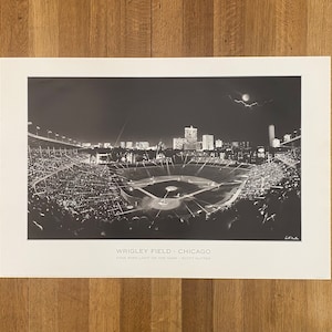 May include: A black and white photograph of Wrigley Field in Chicago at night. The image shows the field, the stands, and the city skyline in the background. The text "Wrigley Field - Chicago" and "Five Star Light on the Game - Scott Hutter" is printed below the image.
