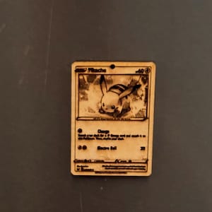 Könnte beinhalten: Pikachu Pokémon-Karte aus Holz mit eingravierten Details. Die Karte zeigt eine Pikachu-Illustration, Text und ein kleines Loch oben. Die Karte ist aus hellfarbenem Holz und hat eine rechteckige Form.