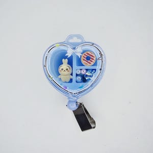 May include: A heart-shaped light blue badge reel featuring a clear front cover. Inside are three compartments containing a small cream-coloured rabbit figurine, a miniature striped doughnut, and several small blue and white spherical beads.