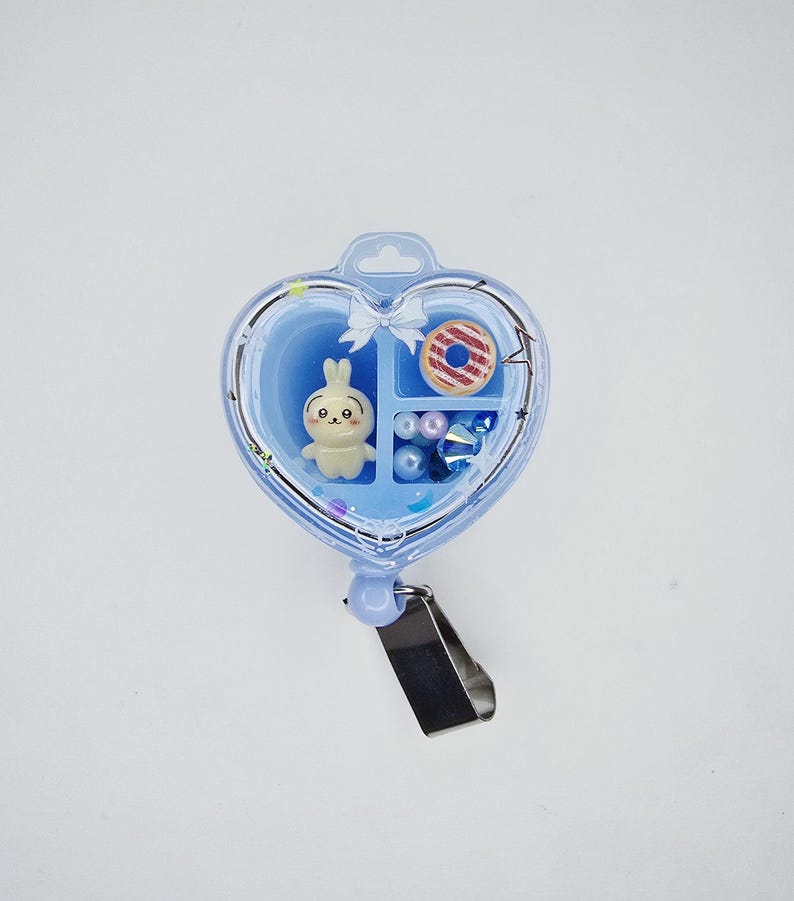 May include: A heart-shaped light blue badge reel featuring a clear front cover. Inside are three compartments containing a small cream-coloured rabbit figurine, a miniature striped doughnut, and several small blue and white spherical beads.