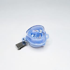 May include: A light blue heart-shaped badge reel with a clear cover containing small decorative charms, including a white bear, a silver bow, and star shapes. The badge reel features a metal clip attached to the side for secure garment attachment.