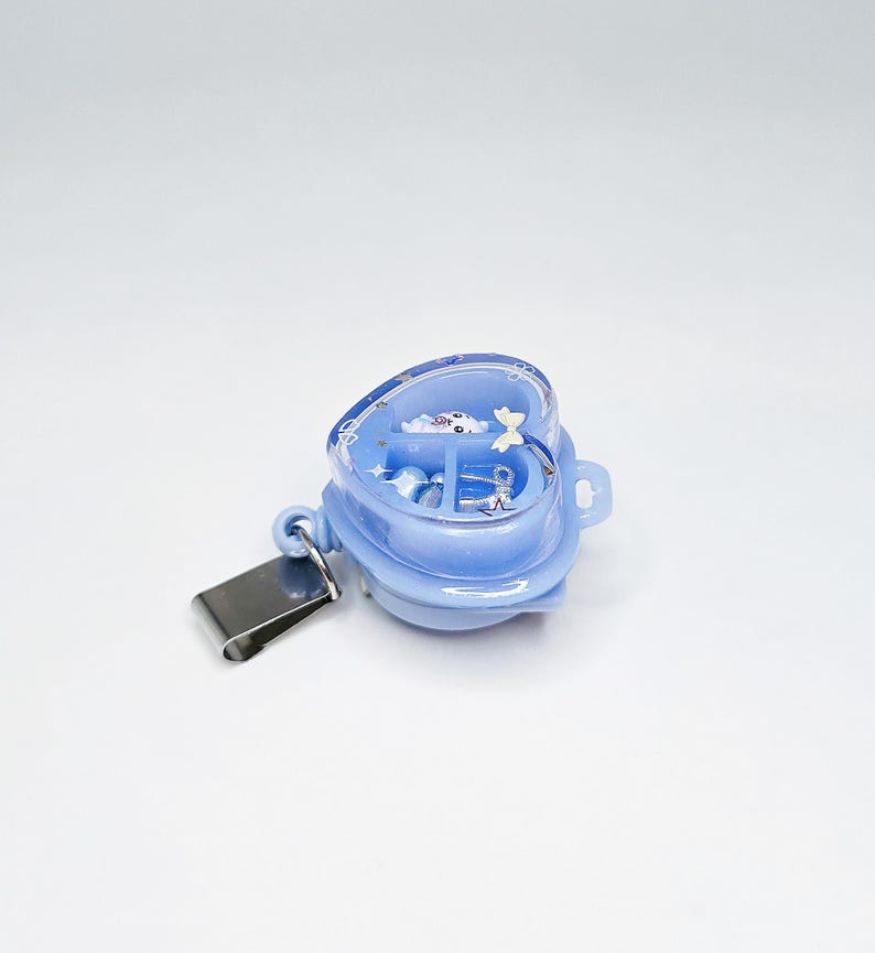 May include: A light blue heart-shaped badge reel with a clear cover containing small decorative charms, including a white bear, a silver bow, and star shapes. The badge reel features a metal clip attached to the side for secure garment attachment.