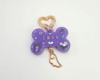 May include: Purple bone-shaped keychain with a gold heart-shaped clasp, a gold angel wing charm, and the name "IRIS" written in gold lettering. The keychain is decorated with glitter, stars, flowers, and a cartoon character.
