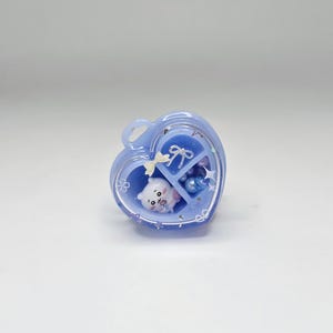 May include: A heart-shaped light blue resin container with a clear lid, featuring internal compartments. Inside are a small white bear figurine, a silver bow charm, a cream-coloured bow, and various blue and purple spherical beads.