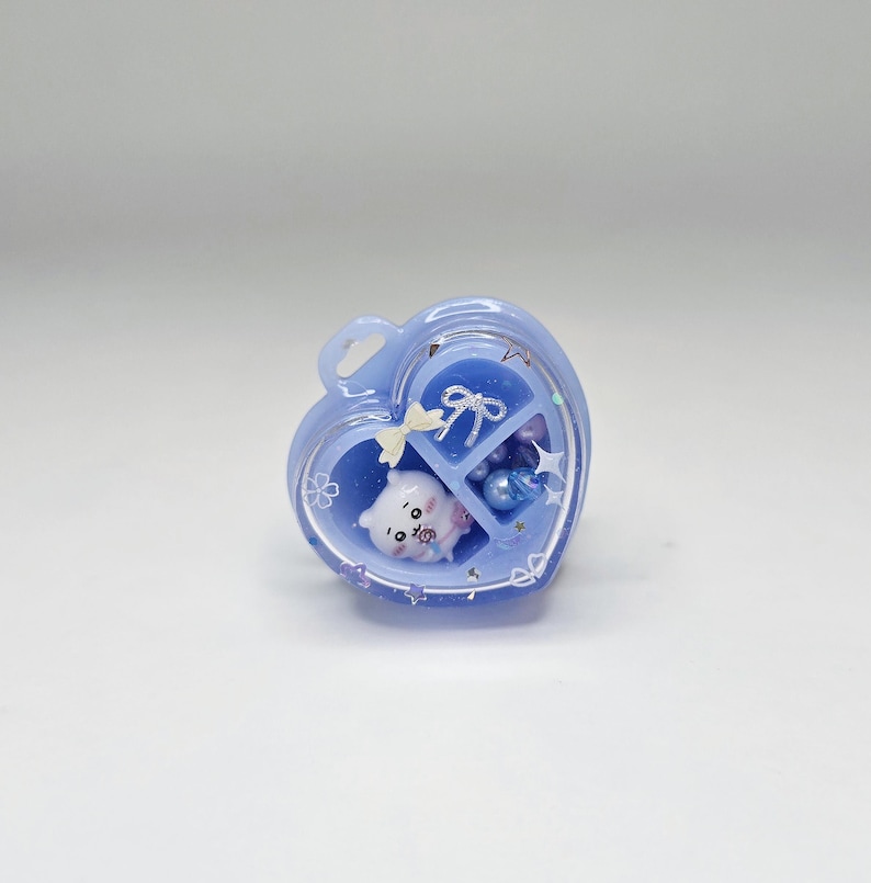 May include: A heart-shaped light blue resin container with a clear lid, featuring internal compartments. Inside are a small white bear figurine, a silver bow charm, a cream-coloured bow, and various blue and purple spherical beads.