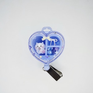 May include: A heart-shaped light purple badge reel featuring a clear front cover with white star and cherry blossom graphics. Inside are three compartments containing a small white cartoon character, a white bow, and several purple beads.