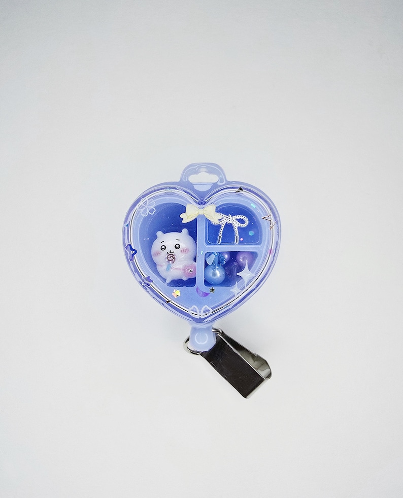 May include: A heart-shaped light purple badge reel featuring a clear front cover with white star and cherry blossom graphics. Inside are three compartments containing a small white cartoon character, a white bow, and several purple beads.