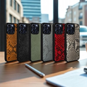 May include: Six smartphone cases with a snakeskin design in shades of orange, black, olive green, grey, red, and tan. Each case has a black border. They are displayed on a wooden surface near a window, with a pen and notepad.