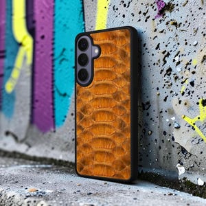 May include: A rectangular smartphone case featuring a textured, orange-brown snakeskin pattern on the back panel. The case has a black frame and a vertical camera module on the upper left side containing three circular lenses and a small flash.