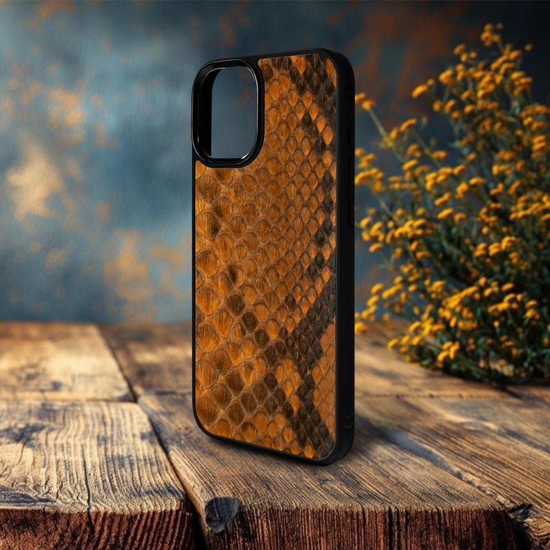 May include: A black phone case with a snakeskin pattern in shades of brown and orange. The case has a sleek design and a protective bumper around the edges. The phone case is displayed on a wooden surface.