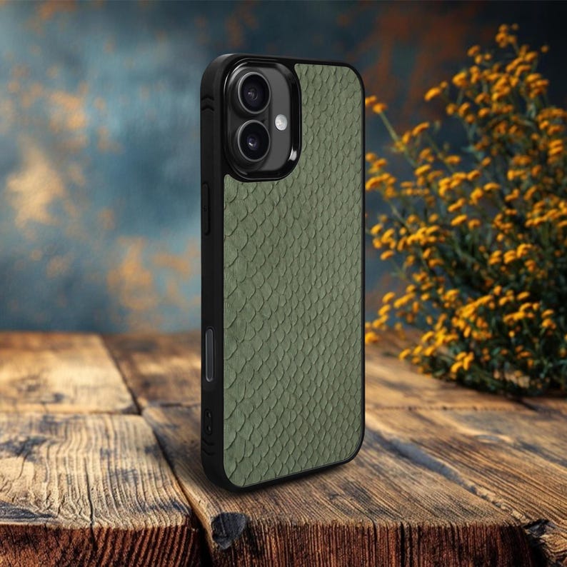 May include: A black phone case with a textured, olive green, snake-skin pattern. The case has a dual-lens camera cutout and is displayed on a wooden surface. The background features a blurred, textured wall and yellow flowers.