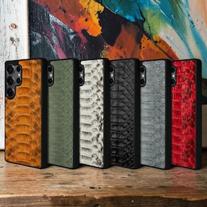 May include: A collection of six rectangular smartphone cases arranged in a row, each featuring a textured snakeskin pattern in distinct colors including orange, olive green, white, black, grey, and red, all encased in a black protective frame.