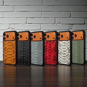 Op de afbeelding: Een verzameling van zes telefoonhoesjes met een slangenhuiddesign. De hoesjes hebben een zwart frame, een oranje camerabehuizing en een verscheidenheid aan kleuren, waaronder goud, zwart, grijs, rood, wit en groen. Ze worden gepresenteerd tegen een witte bakstenen muur.