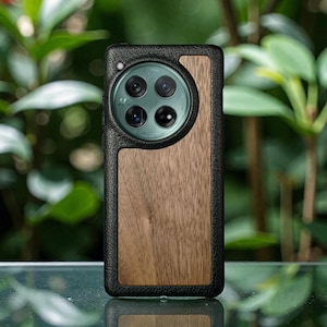 Puede incluir: Una funda para teléfono de cuero negro y madera. La funda presenta un borde de cuero negro, un panel de madera y un recorte circular para la cámara. La funda está diseñada para proteger la cámara y el cuerpo del teléfono.