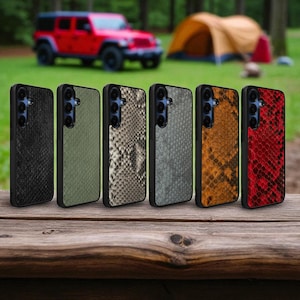 Puede incluir: Seis fundas para teléfono con un diseño de piel de serpiente en varios colores: negro, verde oliva, beige, gris, naranja y rojo. Las fundas se muestran sobre una superficie de madera con un Jeep rojo y una tienda de campaña al fondo.