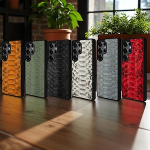 May include: Six phone cases with a snakeskin pattern in shades of orange, green, black, white, grey, and red. Each case has a black frame and a camera aperture.