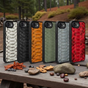 Puede incluir: Seis fundas para teléfono con un patrón de piel de serpiente en varios colores: blanco, negro, naranja, verde, gris y rojo. Cada funda tiene un borde negro y una abertura para la lente de la cámara. Las fundas se muestran sobre una superficie de madera con elementos naturales.