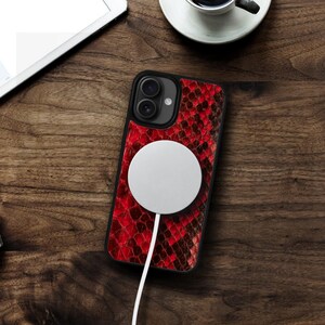 May include: Black iPhone case with a red snakeskin pattern. A white circular charger is attached to the phone with a white cord. The phone and charger are on a wooden surface.