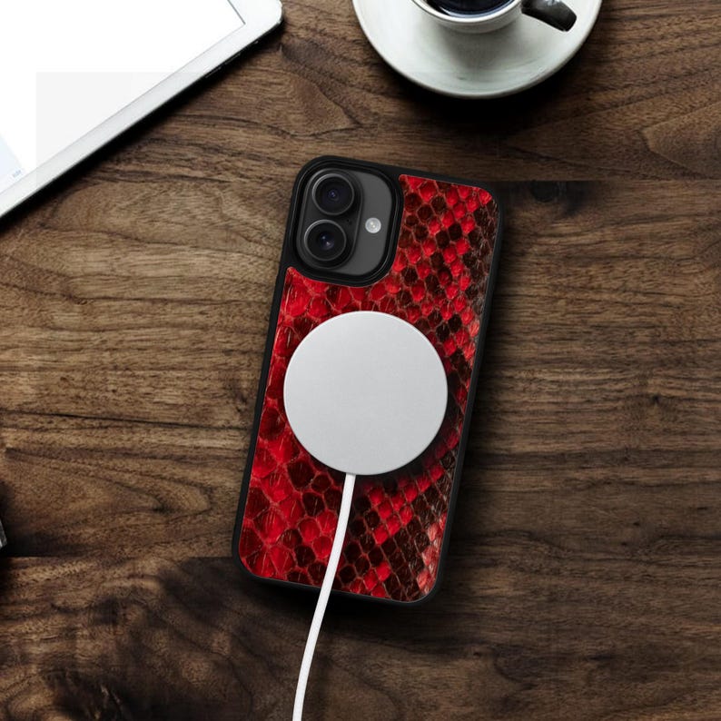 May include: Black iPhone case with a red snakeskin pattern. A white circular charger is attached to the phone with a white cord. The phone and charger are on a wooden surface.