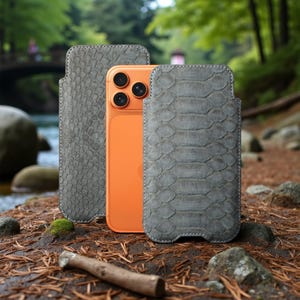 Può includere: Uno smartphone arancione è esposto tra due custodie grigie e strutturate. Le custodie hanno un motivo a pelle di serpente e cuciture bianche. Lo sfondo è un ambiente naturale all'aperto con un ruscello e alberi.