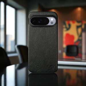 Puede incluir: Una funda de teléfono de cuero negro con una superficie texturizada. La funda tiene un recorte para la cámara en la parte superior y un pequeño logotipo en la parte inferior. La funda está diseñada para un modelo de teléfono específico.