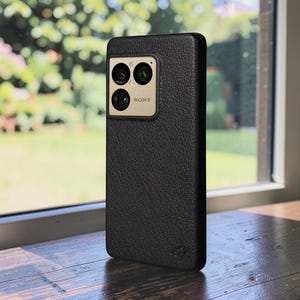 Könnte beinhalten: Eine schwarze Smartphone-Hülle mit einer texturierten, genarbten Oberfläche. Die Hülle verfügt über einen rechteckigen Ausschnitt für das Kameramodul, das drei kreisförmige Linsen und den Schriftzug SONY in Schwarz auf hellgoldem Grund enthält.