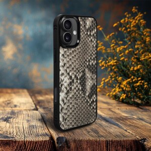 May include: A black phone case with a snakeskin pattern in shades of gray and tan. The case has a dual-lens camera cutout and a sleek, protective design. The case is shown on a wooden surface.