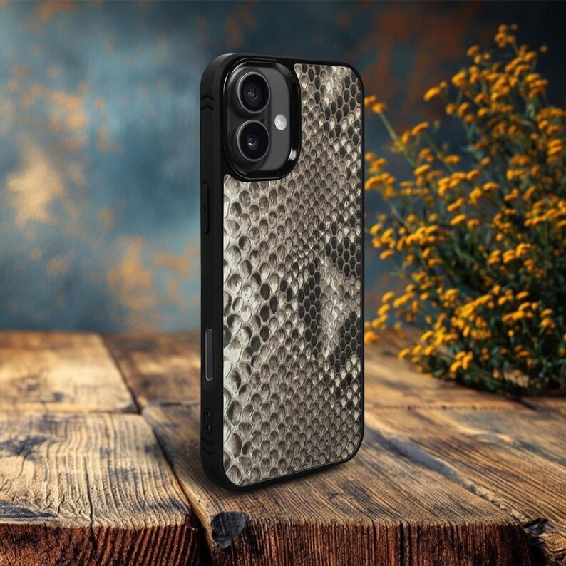 May include: A black phone case with a snakeskin pattern in shades of gray and tan. The case has a dual-lens camera cutout and a sleek, protective design. The case is shown on a wooden surface.