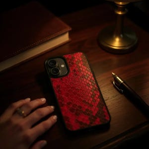 May include: A red and black snakeskin-patterned phone case. The case is on a dark wooden surface, next to a closed brown book, a pen, and a brass lamp. The phone case has a black border and a camera cutout.