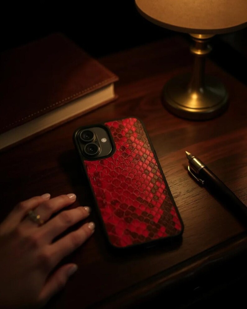 May include: A red and black snakeskin-patterned phone case. The case is on a dark wooden surface, next to a closed brown book, a pen, and a brass lamp. The phone case has a black border and a camera cutout.