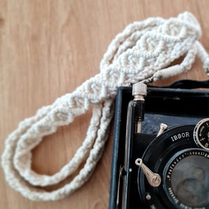 Könnte beinhalten: Eine schwarze Vintage-Kamera mit einem cremefarbenen, geflochtenen Kameragurt. Die Kamera hat silberne Akzente und ein Objektiv mit dem Text "Anastigmat" und "D.R.P." Der Gurt besteht aus weichem, gewebtem Material.